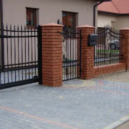 Czarna brama wjazdowa z dekoracyjnymi elementami roślinnymi i furtka z ceglanymi słupkami, widok na posesję z kostki brukowej.